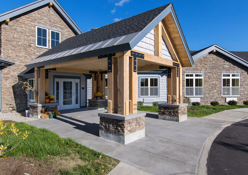 Entrance of a senior living facility with a covered porch supported by wooden beams and stone bases. The building exterior features stone and siding with multiple windows, and there is a concrete driveway and walkway leading to the double glass doors. The sky is clear and blue.