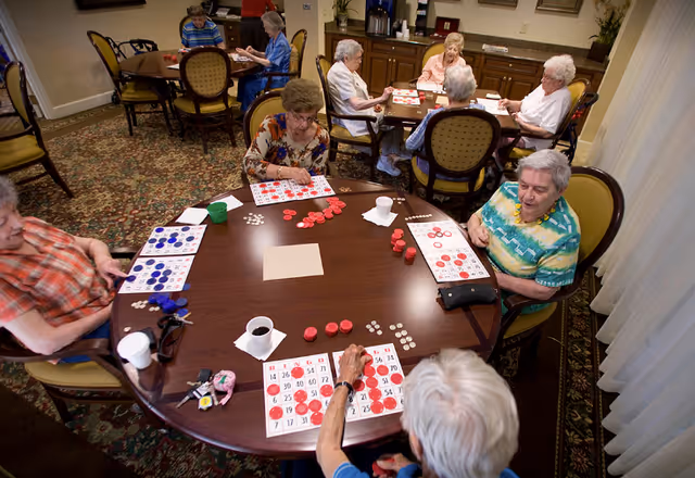 A group of elderly women sitting around two round tables in a common room playing bingo with bingo cards and red and blue chips. The room has patterned carpet, cushioned chairs, and a counter with a coffee machine in the background.