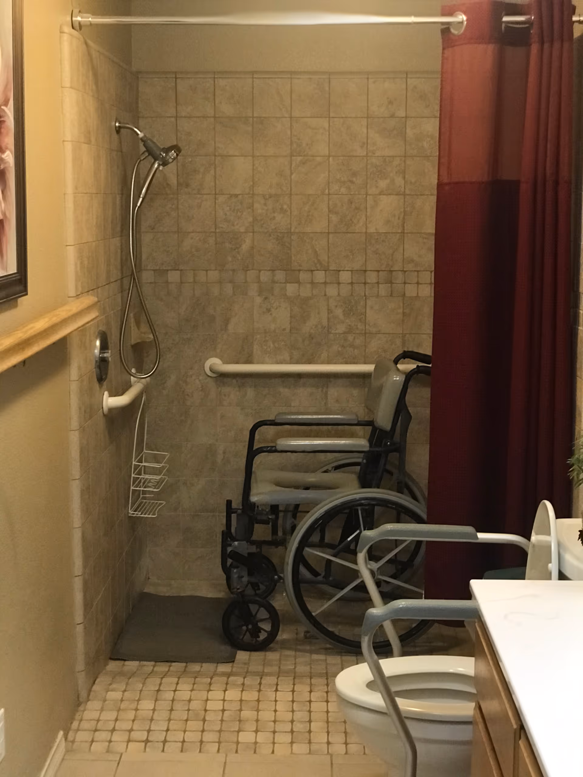 Accessible bathroom with a tiled walk-in shower equipped with a handheld showerhead and grab bars. A wheelchair is positioned inside the shower area. The bathroom also features a toilet with arm supports and a vanity with a sink. A maroon shower curtain is partially drawn to the side.
