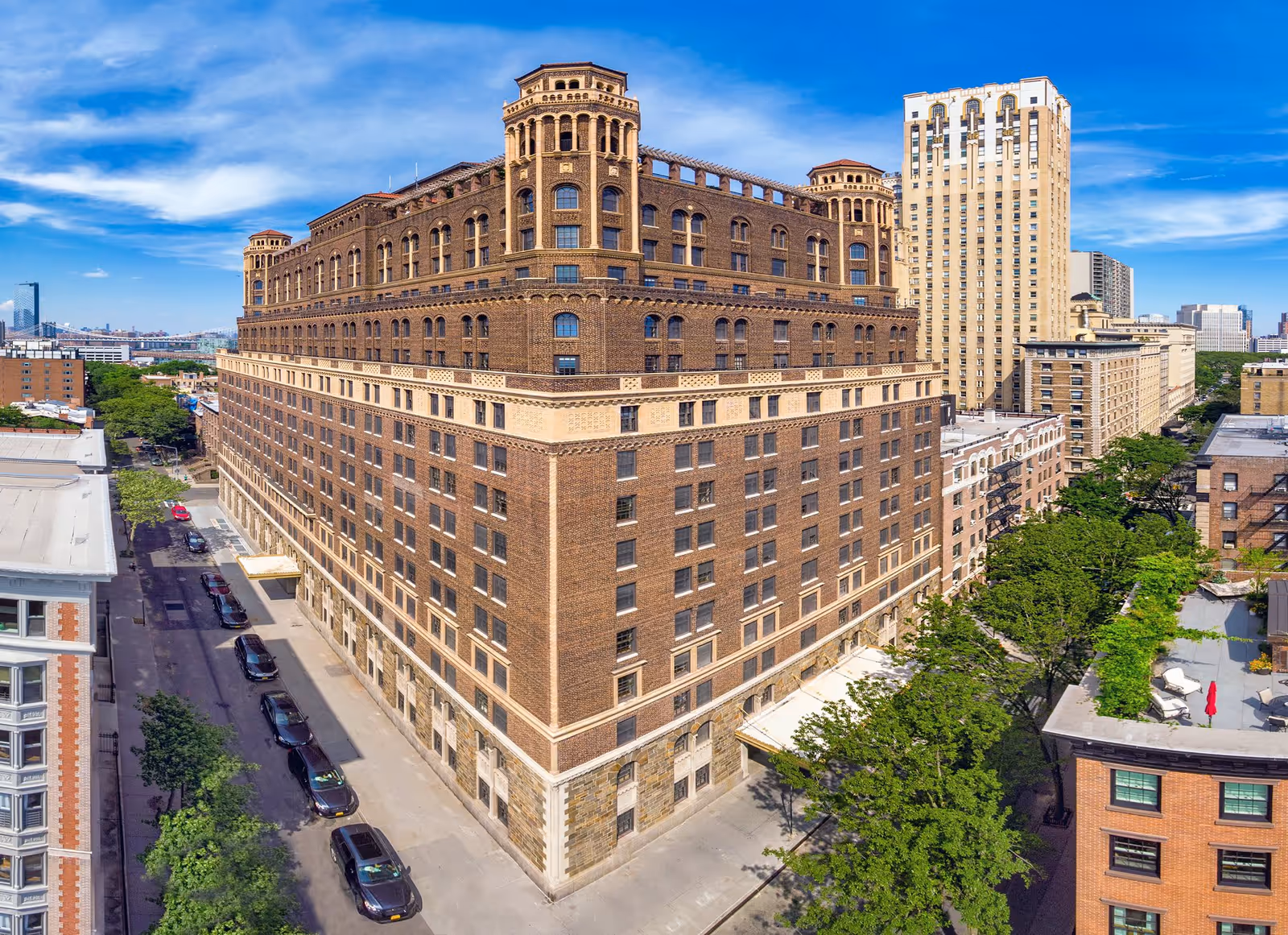 A large, historic multi-story brick building with ornate architectural details, situated on a city street corner with several parked cars and surrounded by other buildings and trees under a blue sky.
