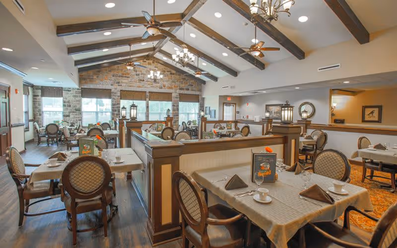 A spacious dining room with multiple tables covered in beige tablecloths, set with cups, saucers, and folded napkins. The room features wooden beams on the ceiling, ceiling fans, chandeliers, and large windows with stone walls letting in natural light. The seating includes cushioned chairs with patterned backs, and the overall decor is warm and inviting.