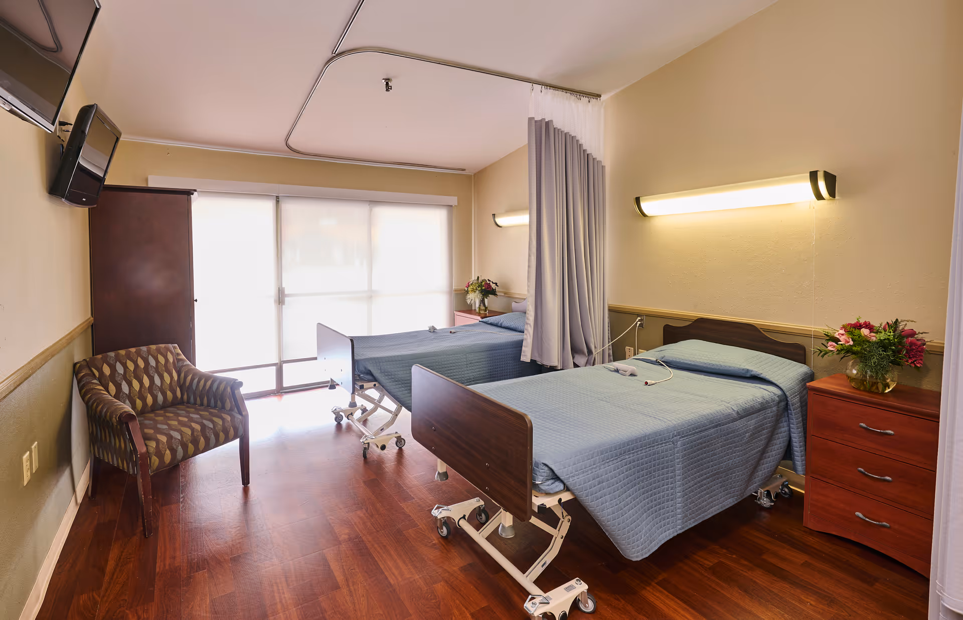 A nursing facility room with two hospital beds covered in blue bedspreads, a wooden nightstand with a vase of flowers, a patterned armchair, a wall-mounted TV, and a large window with blinds letting in natural light. A privacy curtain is partially drawn between the beds.