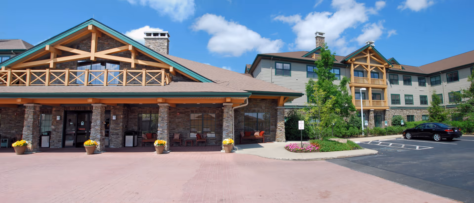 Exterior view of Deer Creek Lodge senior living facility showing a large building with stone and wood accents, a covered entrance with potted flowers, a parking area with a black car, and a clear blue sky with some clouds.