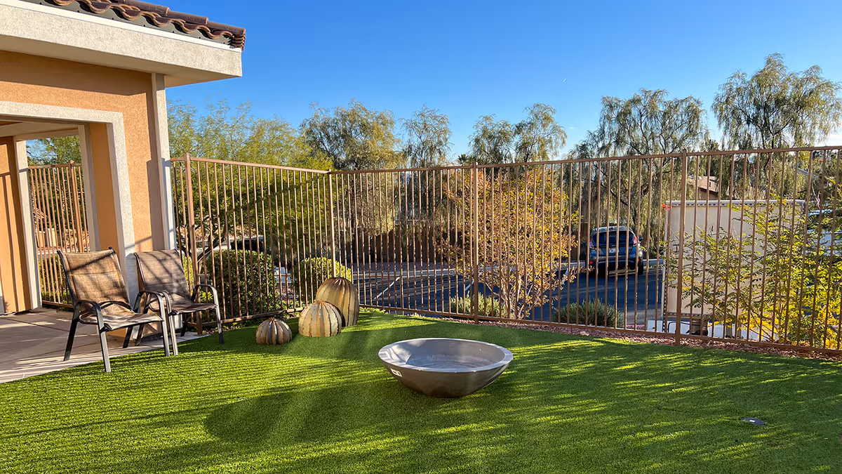 Outdoor patio area with artificial grass, a metal fire pit, three decorative pumpkins, two chairs, and a metal fence overlooking a parking lot and trees under a clear blue sky.