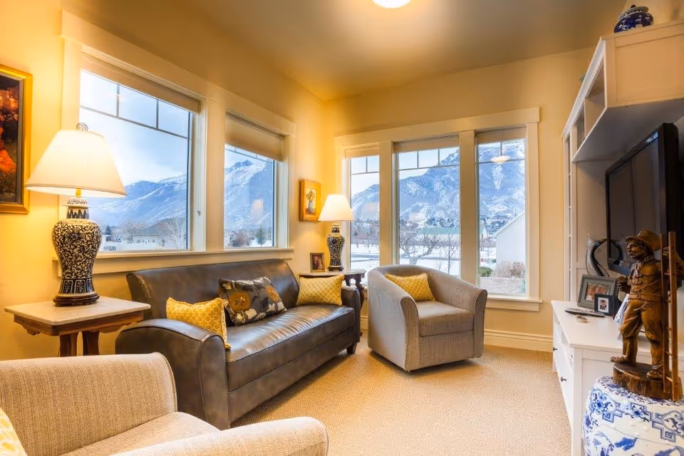 A cozy living room with a dark leather sofa adorned with decorative pillows, two beige armchairs, and two table lamps with blue and white ceramic bases. Large windows reveal a scenic view of snow-covered mountains and a clear sky. A white entertainment unit holds a flat-screen TV and decorative items, including a wooden figurine and framed photos.