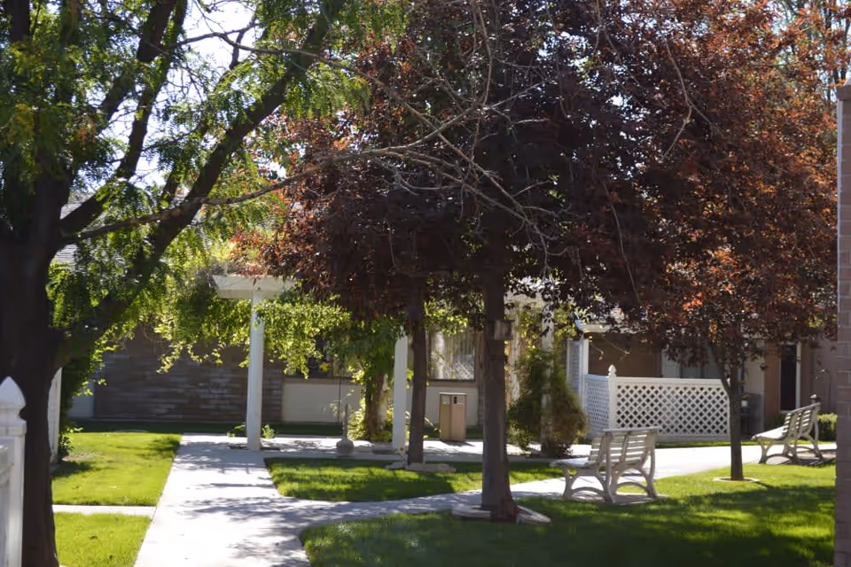 Outdoor area of Truewood by Merrill, Taylorsville featuring a green lawn, several trees with green and reddish leaves, white benches, a paved walkway, and a white lattice fence near a building.