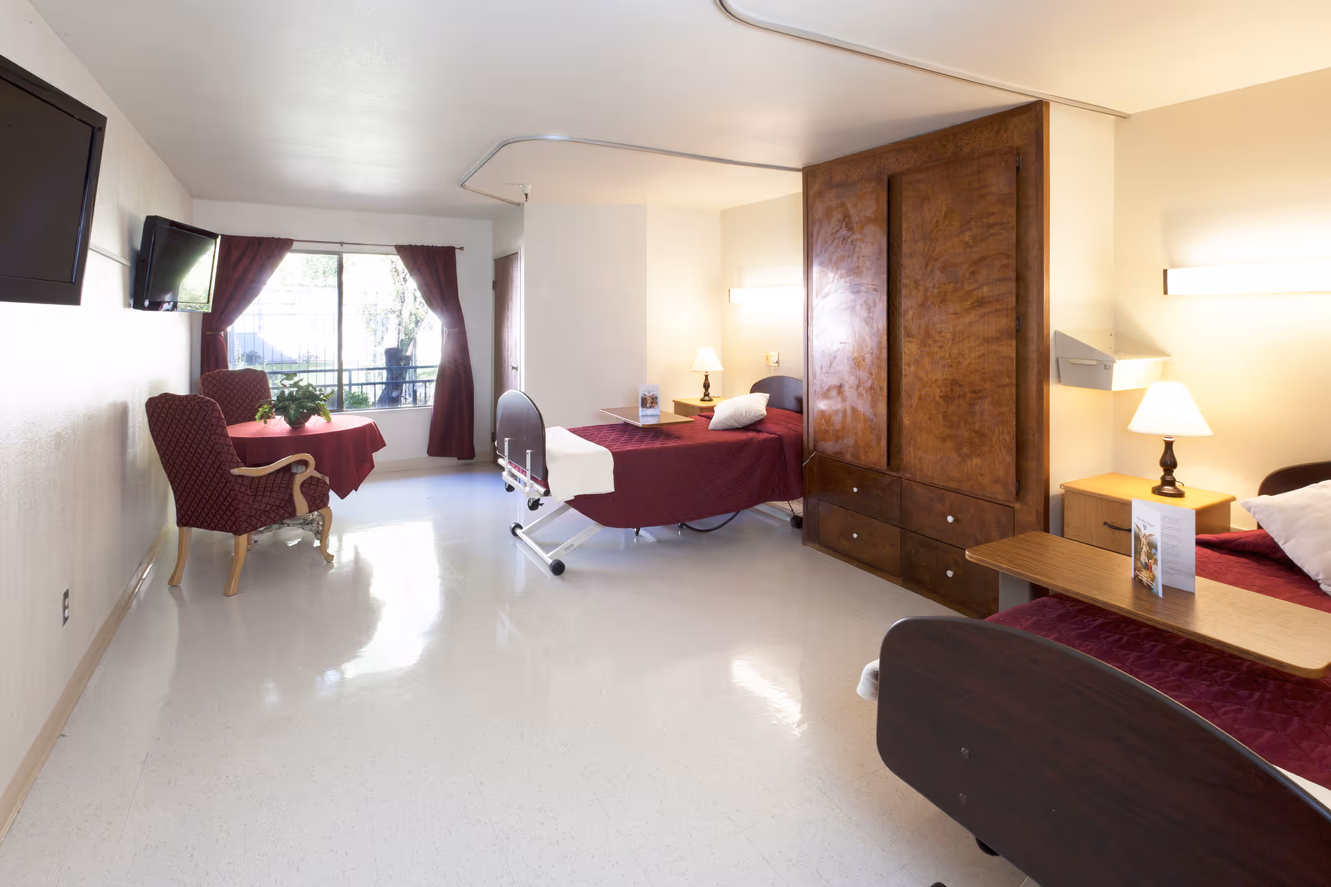 A bright and clean senior living facility bedroom with two hospital-style beds covered in burgundy bedspreads, a wooden wardrobe, bedside tables with lamps, a small table with two upholstered chairs near a window with burgundy curtains, and a wall-mounted television.