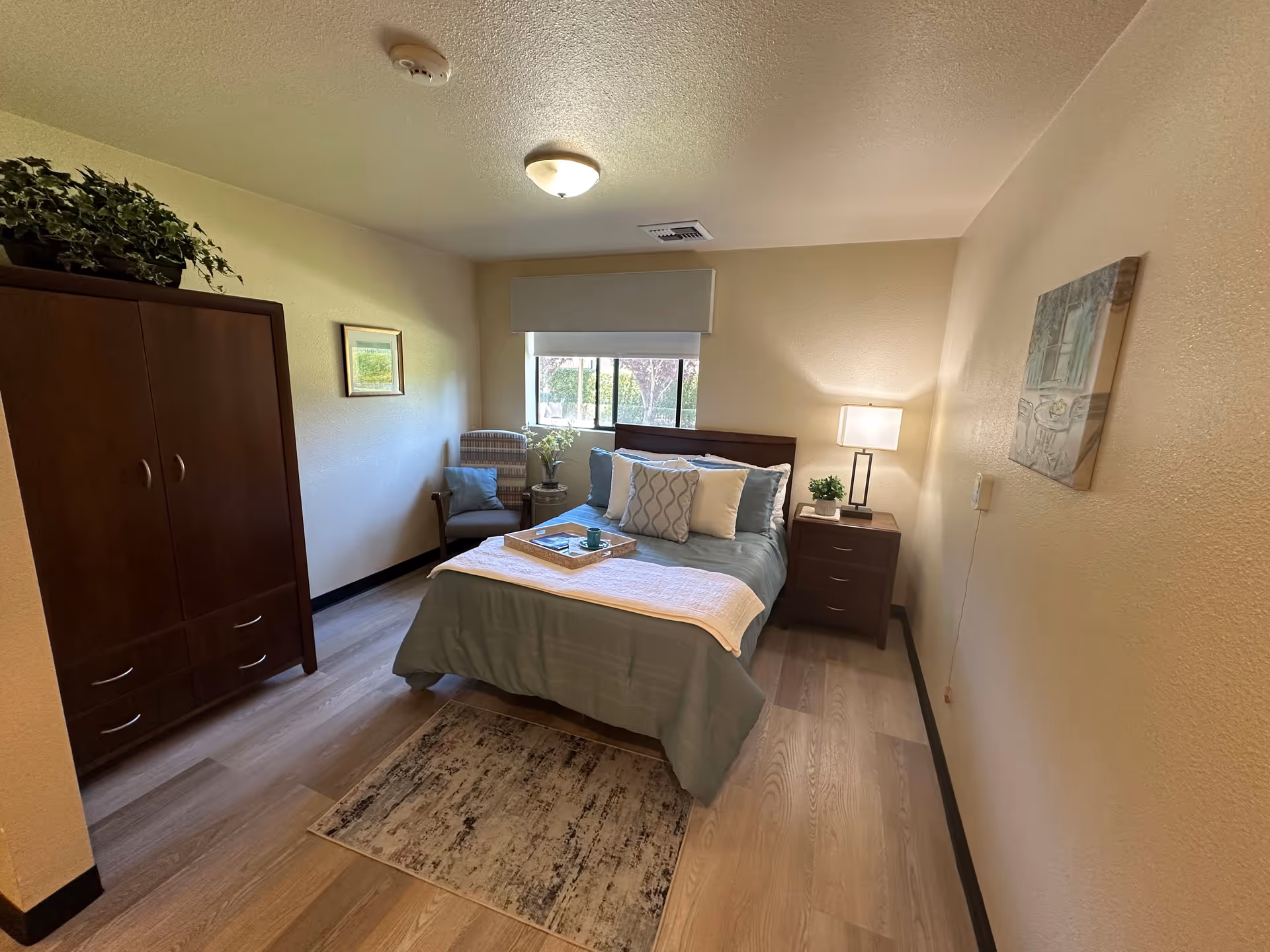 A cozy bedroom with a bed made up with blue and white bedding, a wooden nightstand with a lamp and a small plant, a wooden wardrobe with a green plant on top, a cushioned chair with a pillow, a small side table with flowers, a window with a white valance, and a framed picture on the wall.