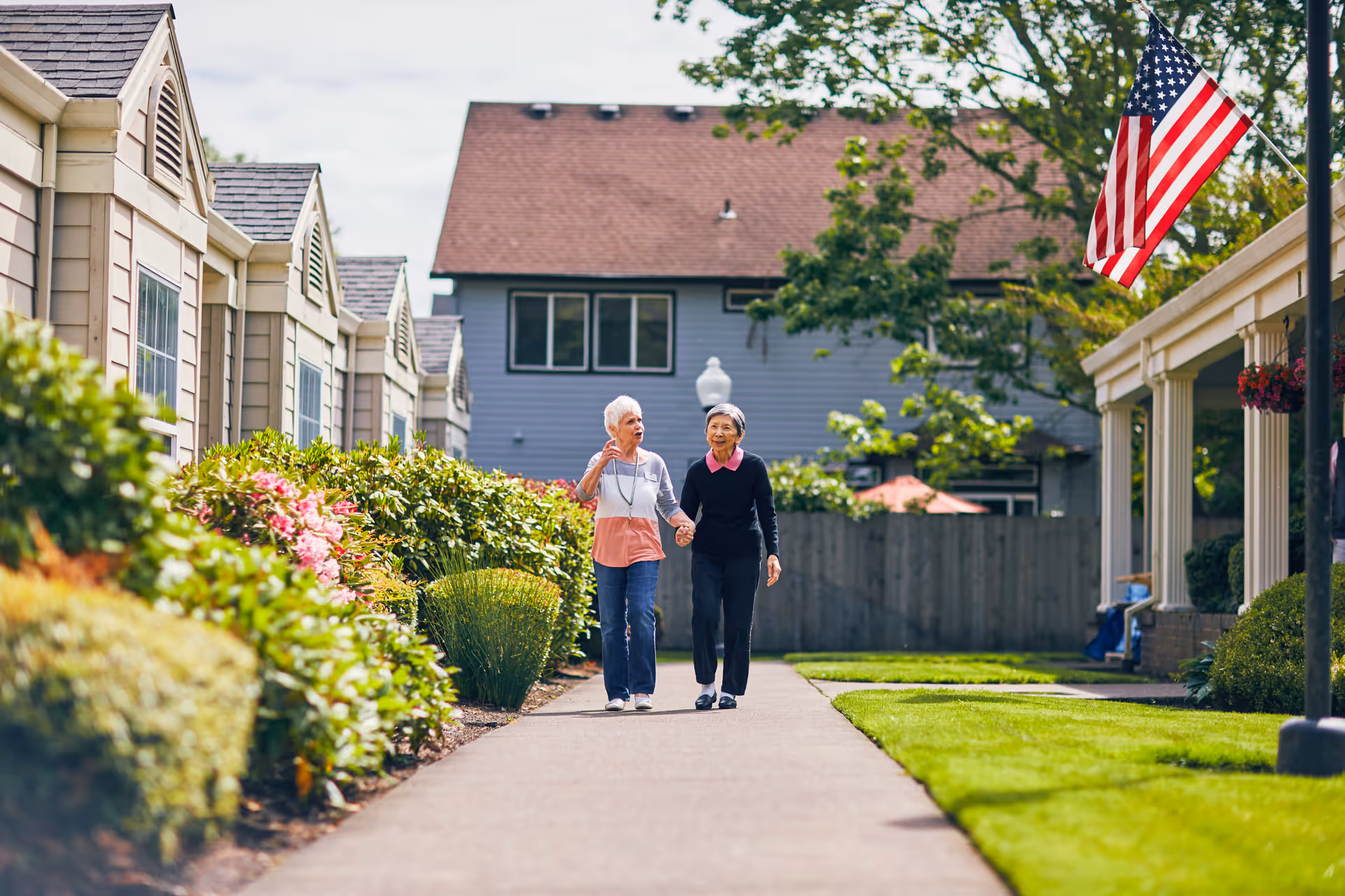 Two elderly women walking hand in hand along a paved pathway in a senior living community with well-maintained gardens and residential buildings on either side. An American flag is visible on a pole to the right.