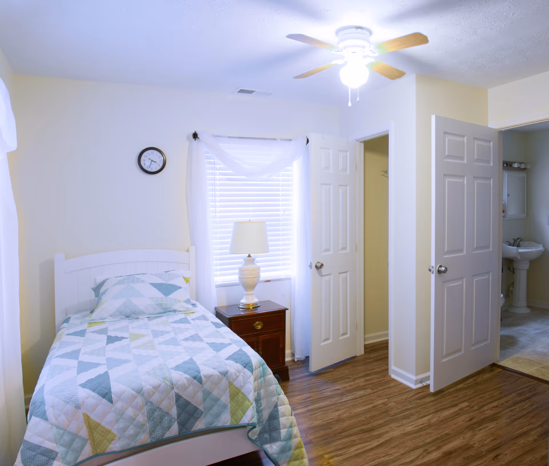 A small bedroom with a single bed covered with a geometric patterned quilt in shades of blue, green, and white. Next to the bed is a wooden nightstand with a white lamp. A window with white blinds and sheer curtains is behind the nightstand. The room has light-colored walls, wood flooring, a ceiling fan with a light, and two open doors leading to a closet and a bathroom with a pedestal sink.