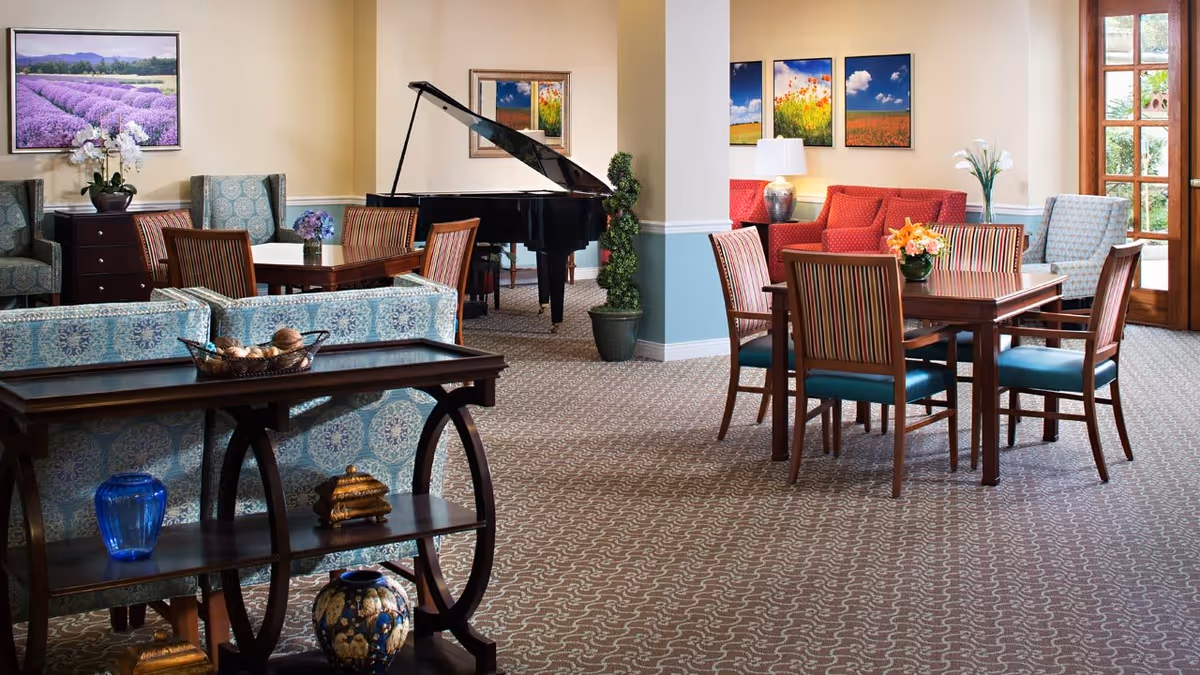 A spacious senior living common area with patterned carpet, multiple seating arrangements including upholstered chairs and sofas, wooden tables with floral centerpieces, a black grand piano, and colorful landscape paintings on the walls. Large windows and a glass door allow natural light into the room.