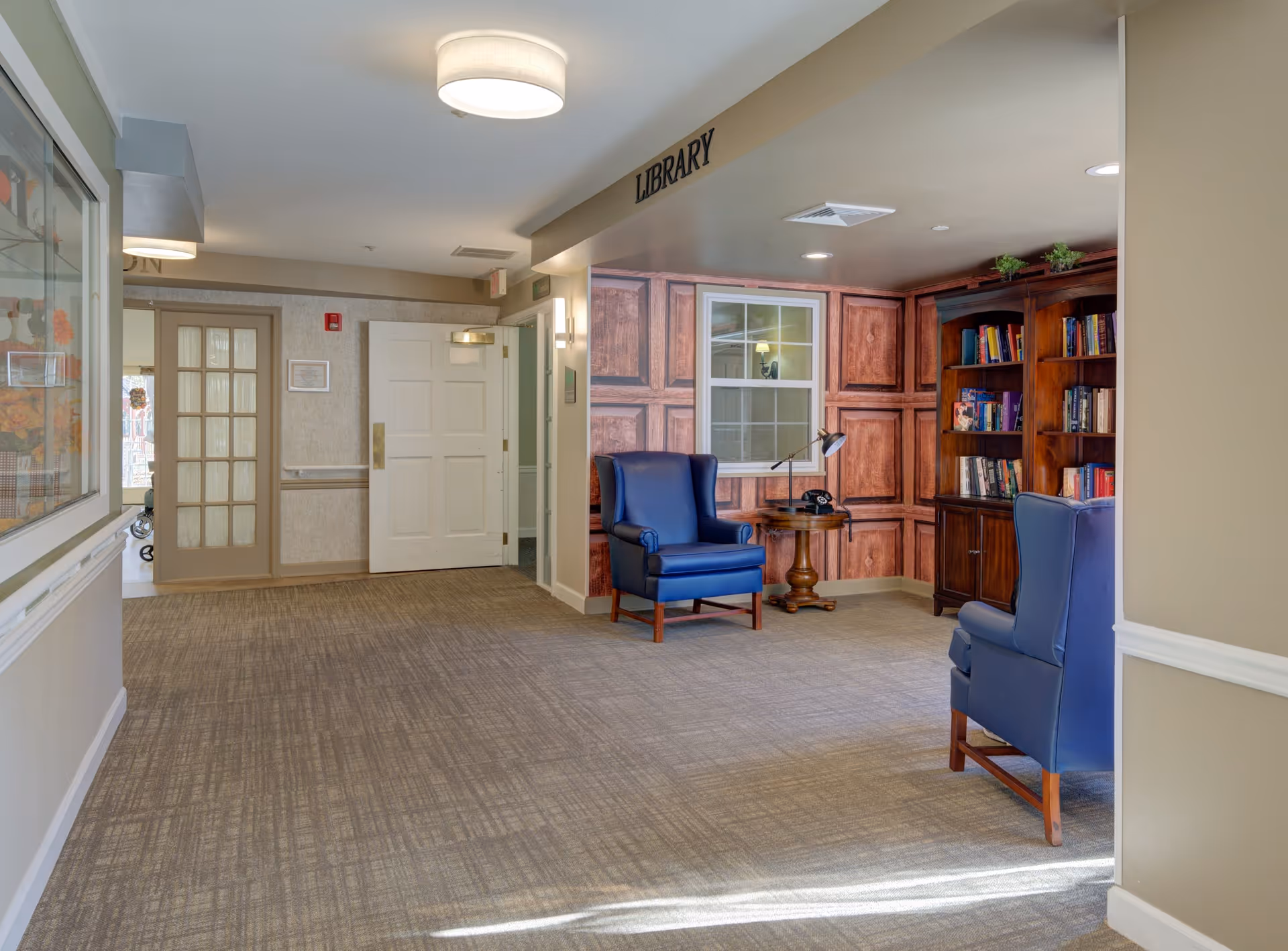 Interior view of a quiet library area in a senior living facility. The space features two blue armchairs, a small wooden side table with a lamp and a vintage telephone, and a wooden bookshelf filled with books. The walls have a wood panel design, and there is a window above the side table. The carpeted floor and soft lighting create a cozy atmosphere. A sign above the area reads 'LIBRARY'.