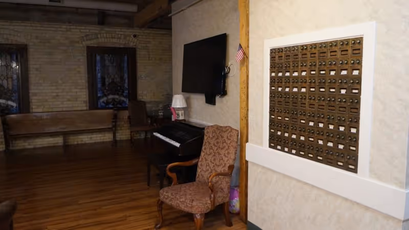 Interior room with wooden floor and brick walls featuring stained glass windows. There is a wooden bench, a patterned armchair, an electronic keyboard on a stand, a wall-mounted flat screen TV, and a wall-mounted set of small mailboxes. A small American flag is attached to a wooden post near the mailboxes.