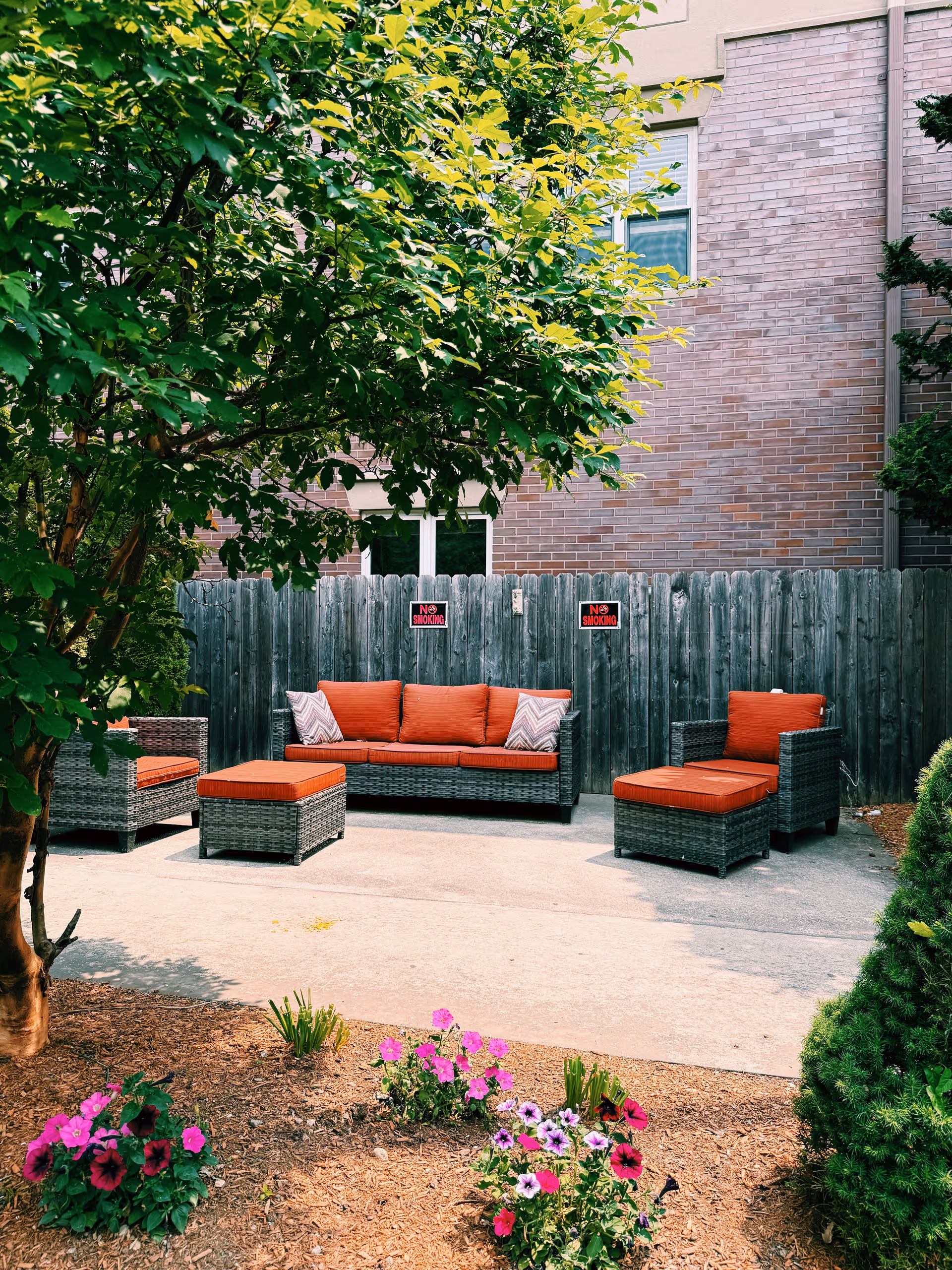 Patio area with wicker sofas and red cushions against a wooden fence and brick building, framed by a tree and flowering plants.