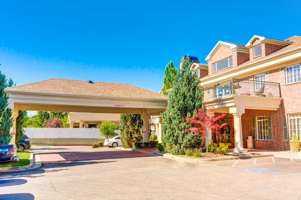 Exterior view of a senior living facility named Escalante at Coventry, showing a brick building with a covered entrance, balcony, and surrounding greenery including trees and shrubs under a clear blue sky.
