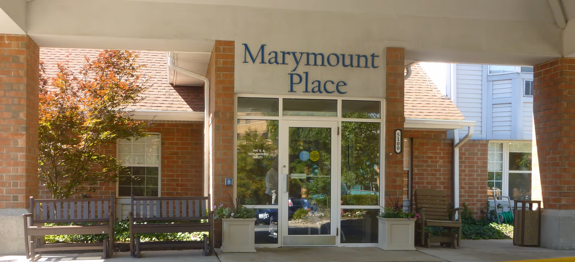 Entrance to Marymount Place Assisted Living facility with glass double doors, two wooden benches, a wooden chair, potted plants, and a tree with autumn-colored leaves. The building exterior features brick and siding.