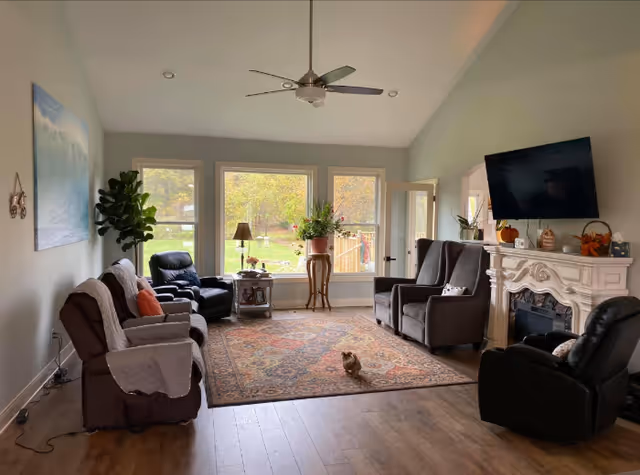 Sunlit communal living room with several recliner chairs around a patterned rug, large windows overlooking a yard, a wall-mounted TV above a mantel, a ceiling fan, and a small pet on the rug.