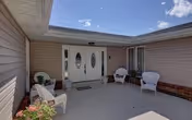 Outdoor patio area at Dream Home Assisted Living with beige siding walls, two white double doors with oval glass panels, four white chairs arranged around the patio, potted plants, and a clear blue sky above.