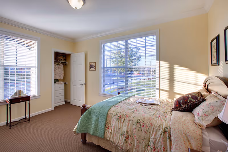 A bright bedroom with beige walls and carpet, featuring a bed with floral bedding and a green throw blanket. There are two large windows with white blinds letting in natural light, a small wooden side table with a decorative item, and an open closet with clothes hanging inside. The room has framed artwork on the walls and a ceiling light fixture.