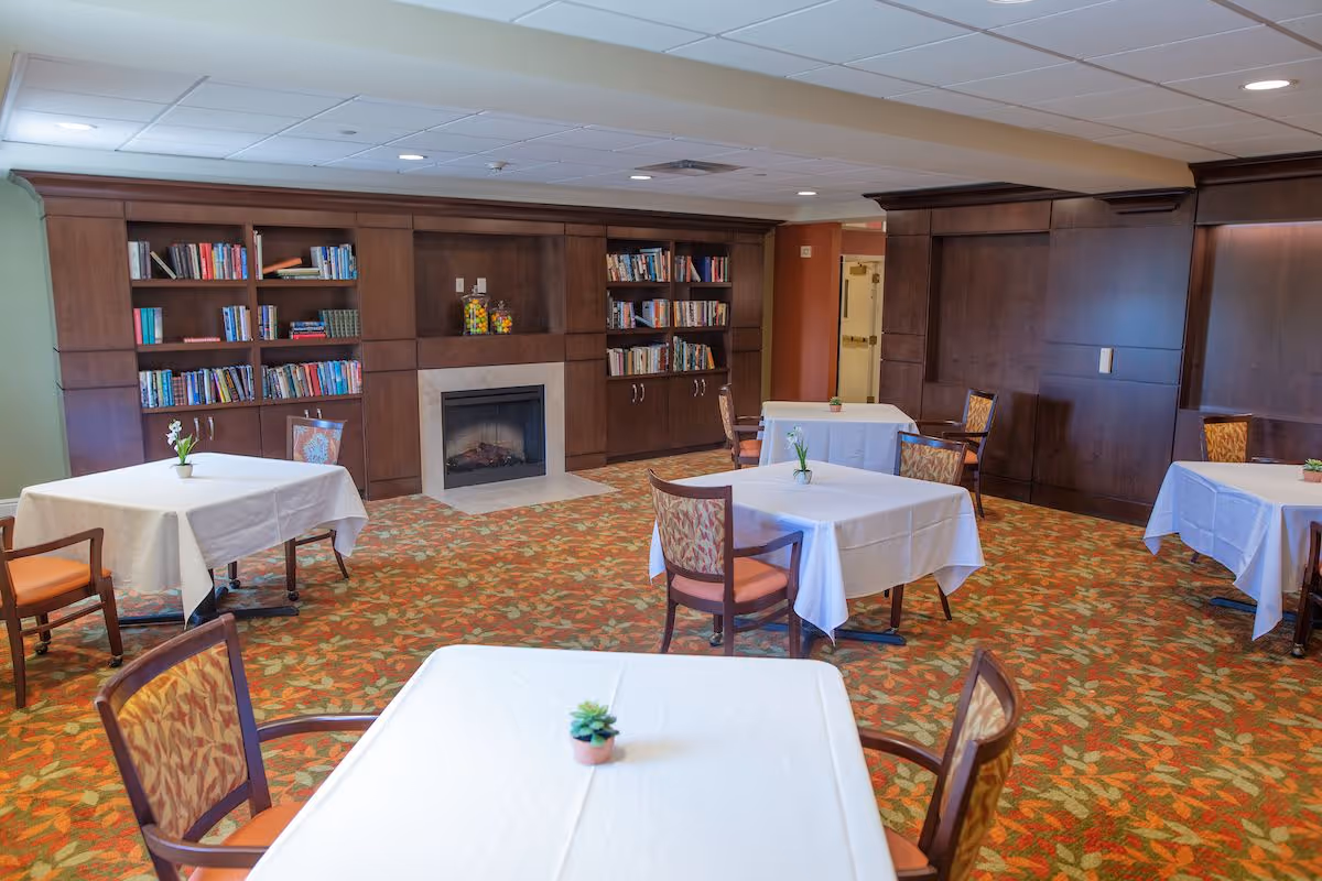 A cozy common area with several tables covered in white tablecloths, each with a small potted plant. The room features a patterned carpet with autumn colors, wooden chairs with patterned cushions, a built-in wooden bookshelf filled with books, and a fireplace in the center of the bookshelf unit. The ceiling has recessed lighting.