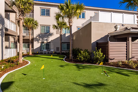 Outdoor putting green area with small yellow flags surrounded by palm trees and landscaping, adjacent to a three-story beige building under a clear blue sky.