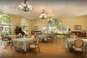 A spacious dining room with multiple round tables covered with green patterned tablecloths, each surrounded by chairs. The room features large arched windows letting in natural light, chandeliers hanging from the ceiling, a piano in the corner, and framed artwork on the walls.