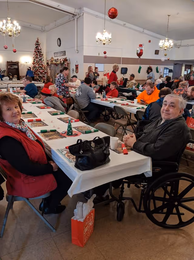 A large room decorated for Christmas with multiple tables where elderly people and caregivers are gathered. The tables are covered with white tablecloths and have Christmas-themed decorations. A decorated Christmas tree and hanging ornaments are visible in the background. People are seated and appear to be enjoying a social event.