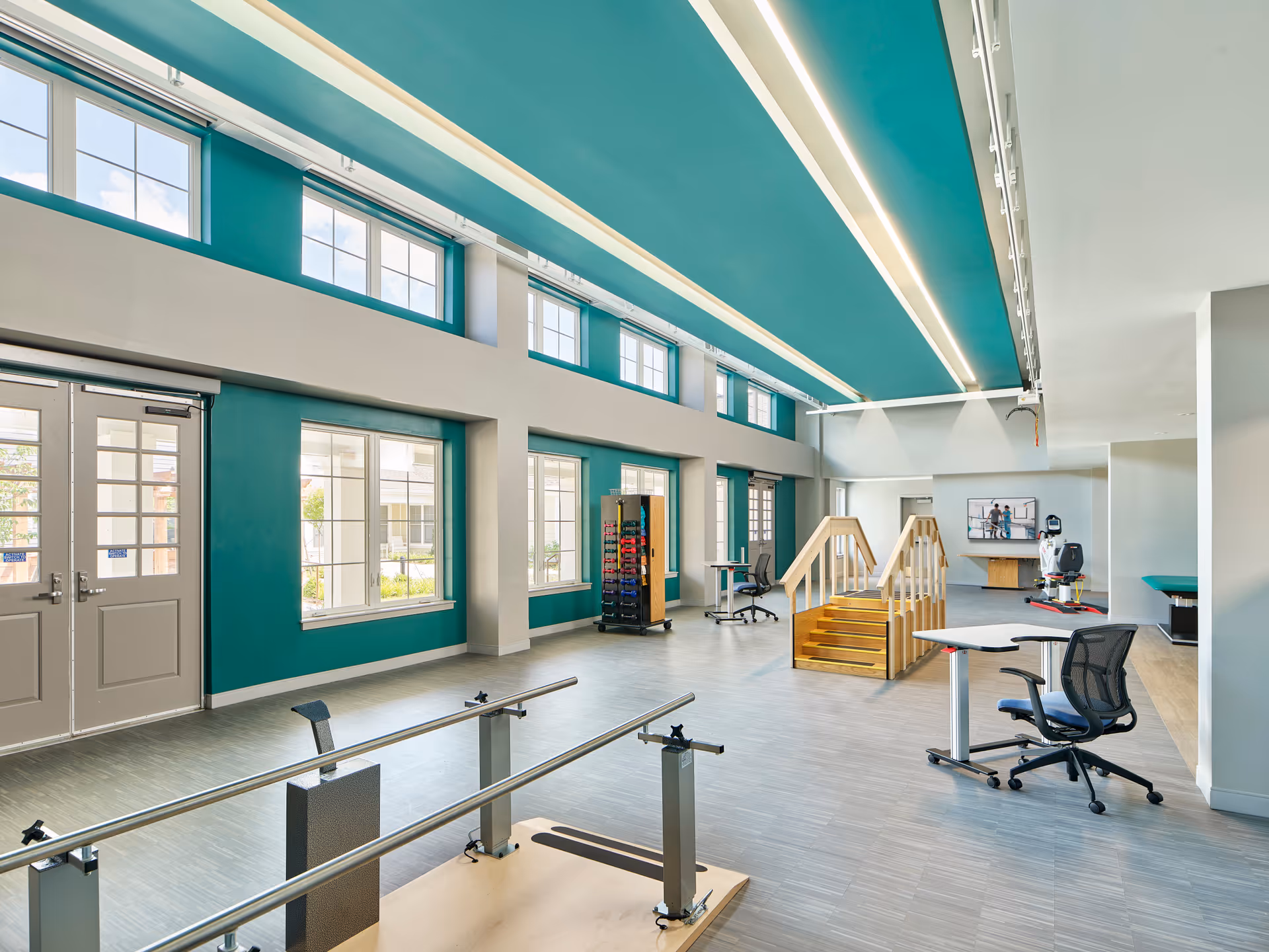 Bright and spacious rehabilitation room with large windows, teal accent walls, parallel bars for walking exercises, a small wooden staircase for physical therapy, office chairs with desks, and exercise equipment in the background.