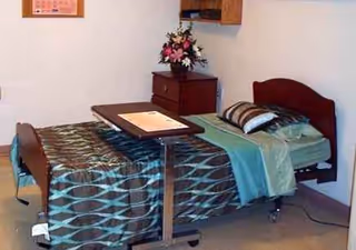 Bedroom in a senior care facility with a single bed, desk, and floral arrangement.