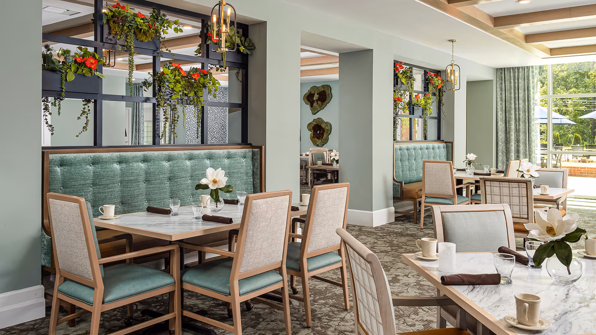 A bright and elegant dining room with multiple tables and chairs arranged neatly. The tables have white marble tops with cups, glasses, and napkins placed on them. There are green cushioned bench seats along the walls, and decorative hanging plants add a touch of nature. Large windows allow natural light to fill the room, and the overall color scheme is soft green and beige.