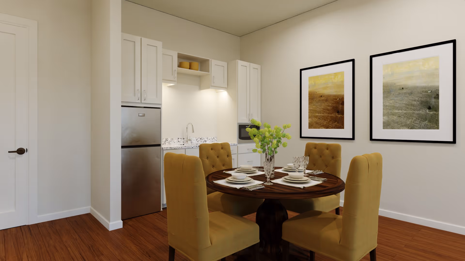 Small dining area with a round table set for four and mustard upholstered chairs beside a compact kitchenette and two framed artworks on the wall.