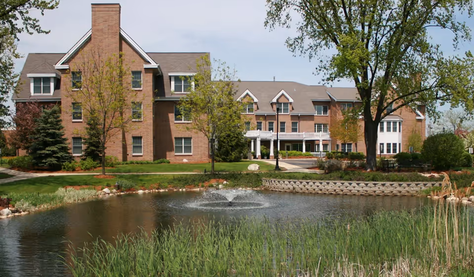 A large brick building with multiple windows and a peaked roof, surrounded by trees and greenery. In front of the building is a pond with a water fountain in the center, bordered by tall grass and landscaping.