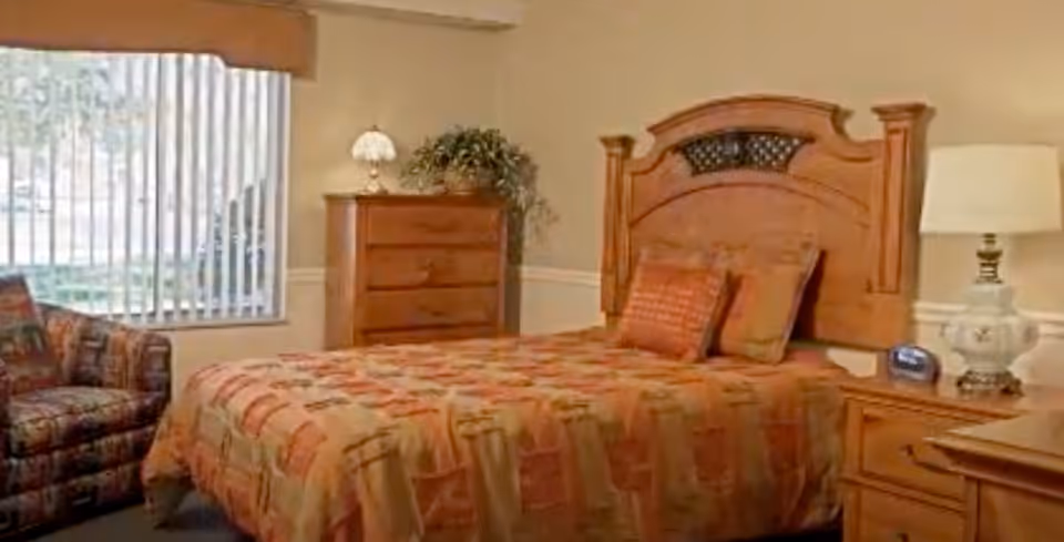 A cozy bedroom with a wooden bed frame and headboard, a bed covered with a patterned orange and beige bedspread, two pillows, a wooden dresser with a lamp and a plant on top, a wooden nightstand with a lamp and a clock, and a patterned armchair near a large window with vertical blinds.