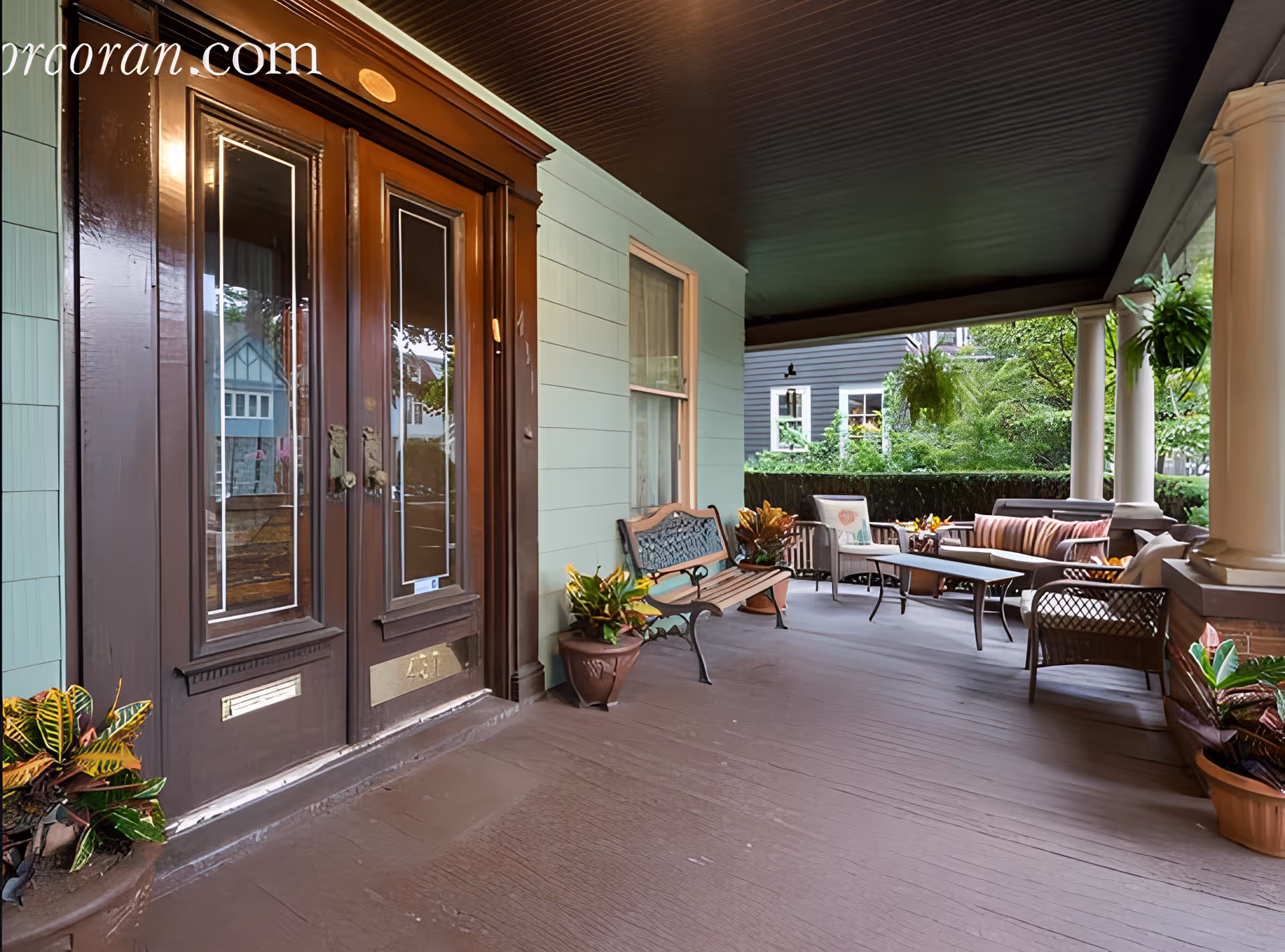 A spacious covered porch area with a dark wooden double door entrance on the left. The porch features a wooden bench with decorative metal backrest, several potted plants, and a seating area with cushioned chairs, a sofa, and a coffee table. The porch ceiling is dark, and white columns support the roof. Greenery and neighboring houses are visible in the background.