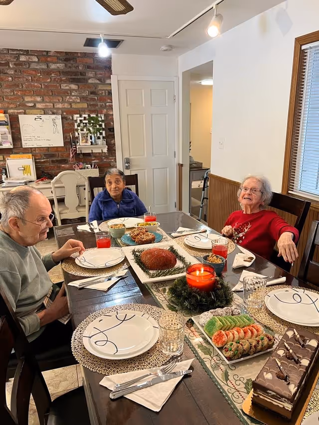 Three elderly people sitting around a dining table set with plates, glasses, and various desserts including cookies, cake, and a glazed ham centerpiece. The room has a brick wall, a white door, and a window with blinds.