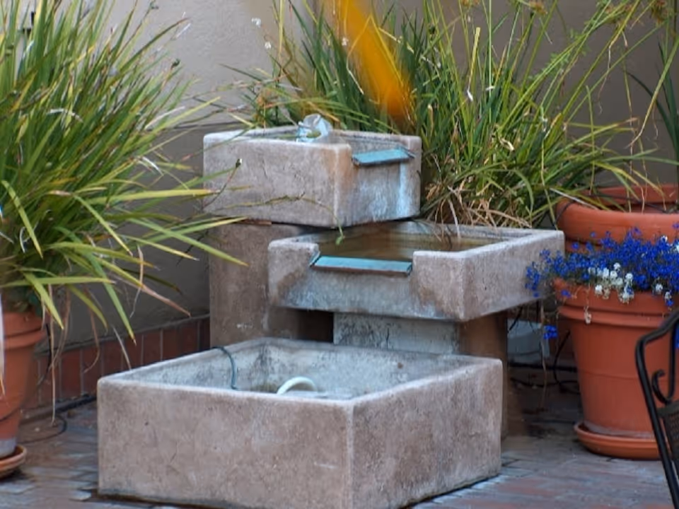 A tiered stone water fountain surrounded by potted plants with green foliage and small blue and white flowers, set on a brick patio against a beige wall.