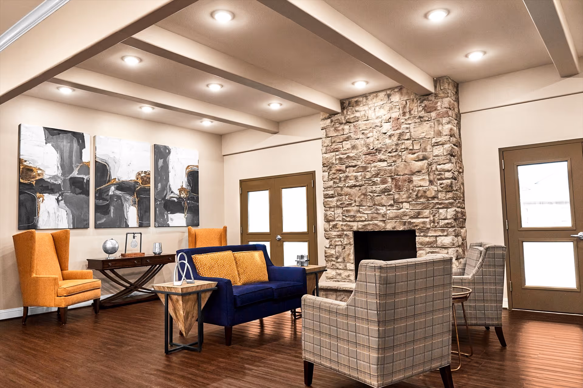 A cozy living room area with a stone fireplace, two plaid armchairs, a blue sofa with orange cushions, two orange high-back chairs, a wooden side table, and abstract artwork on the wall. The room has wooden flooring, beige walls, and recessed ceiling lights.