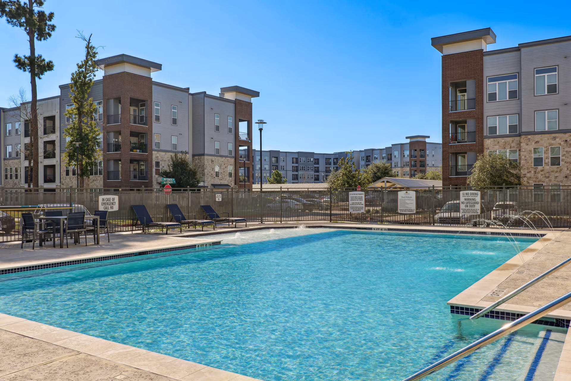 Outdoor swimming pool with clear blue water surrounded by lounge chairs and tables. In the background, there are multi-story residential buildings under a clear blue sky.