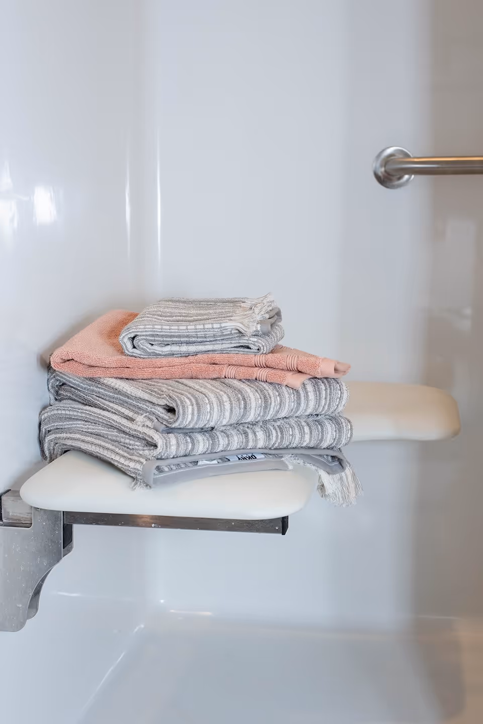 Folded towels in shades of gray and pink placed on a white fold-down shower seat inside a white shower stall with a metal grab bar.