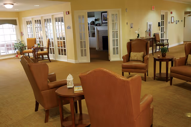 A spacious and well-lit common area in a senior living facility with several comfortable armchairs arranged around small wooden tables. The room features beige walls, carpeted floors, and large windows with white shutters allowing natural light to enter. In the background, there are French doors leading to another room with a fireplace and built-in shelves.