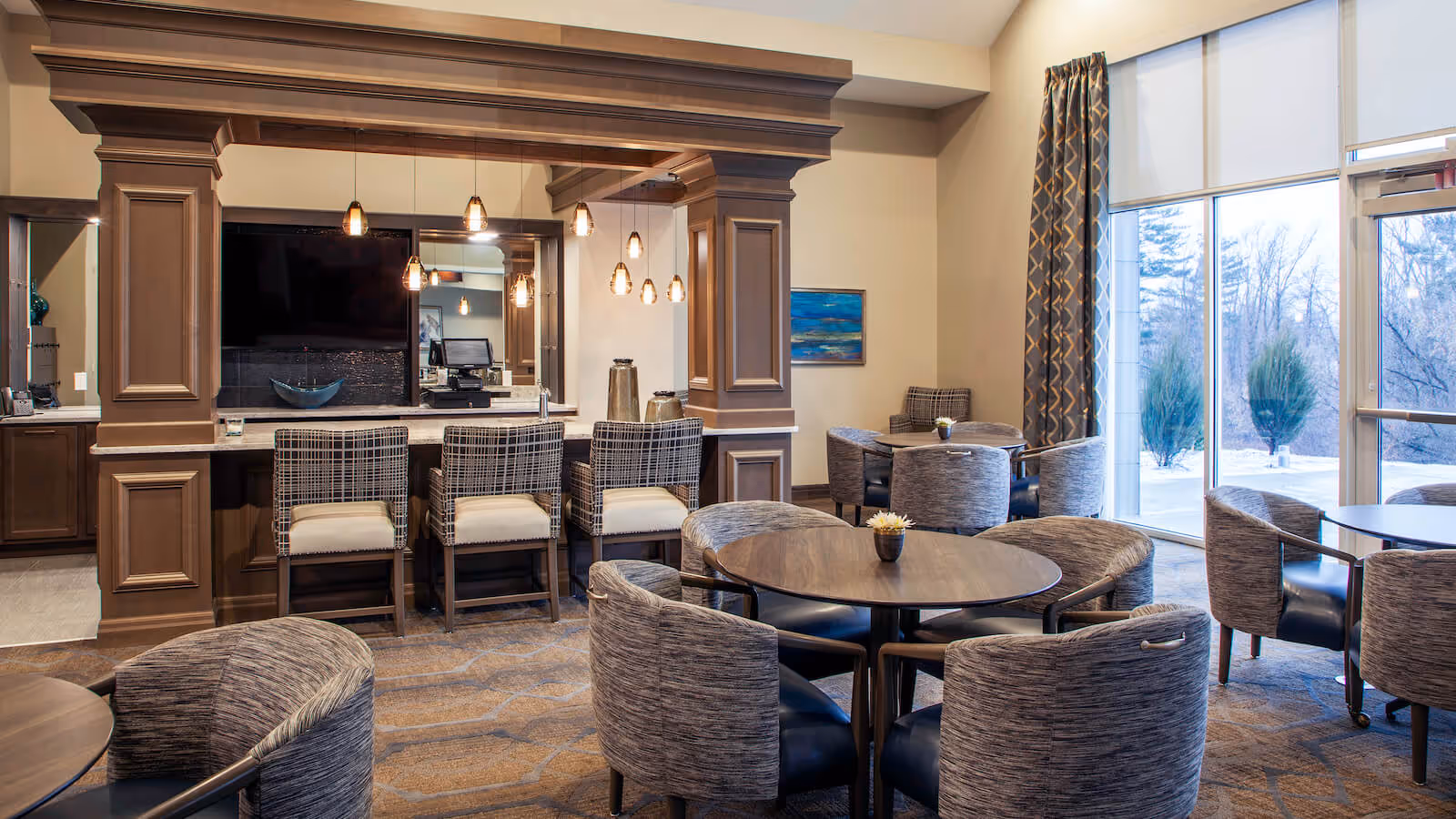 A cozy and modern common area with round tables surrounded by cushioned chairs, a bar counter with high chairs, pendant lights hanging above, large windows with patterned curtains letting in natural light, and a view of trees outside.