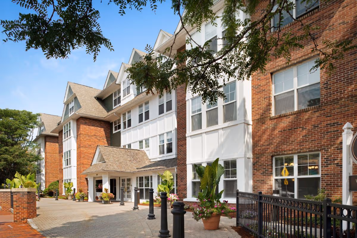 Exterior view of a multi-story brick and white panel building with large windows, a covered entrance, potted plants, and a paved walkway under a blue sky with some tree branches overhead.