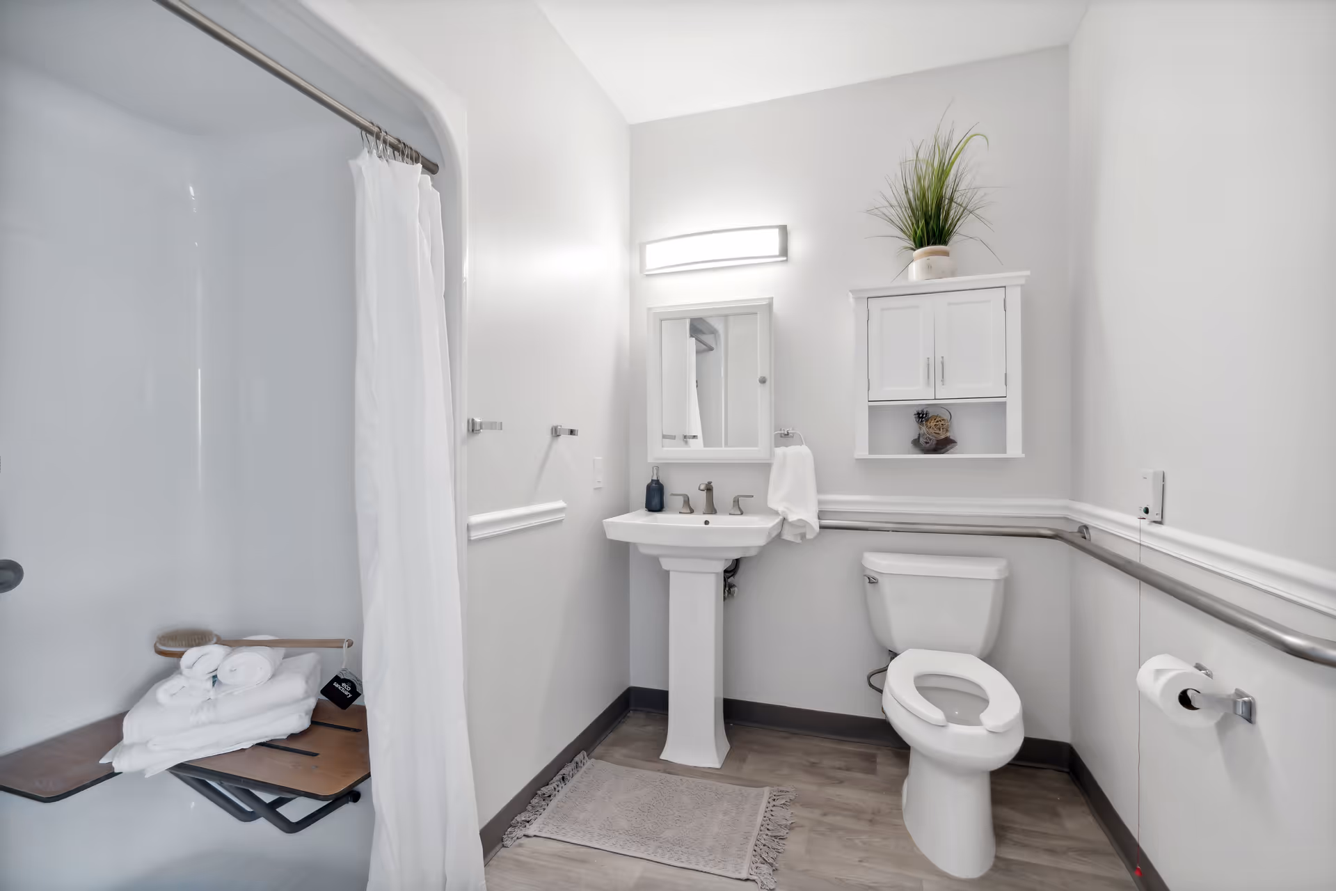 A clean and accessible bathroom with a white pedestal sink, a toilet with grab bars on both sides, a shower with a white curtain and a fold-down seat holding towels and a brush, a wall-mounted cabinet with a plant and decorative items, and a light fixture above a mirror.