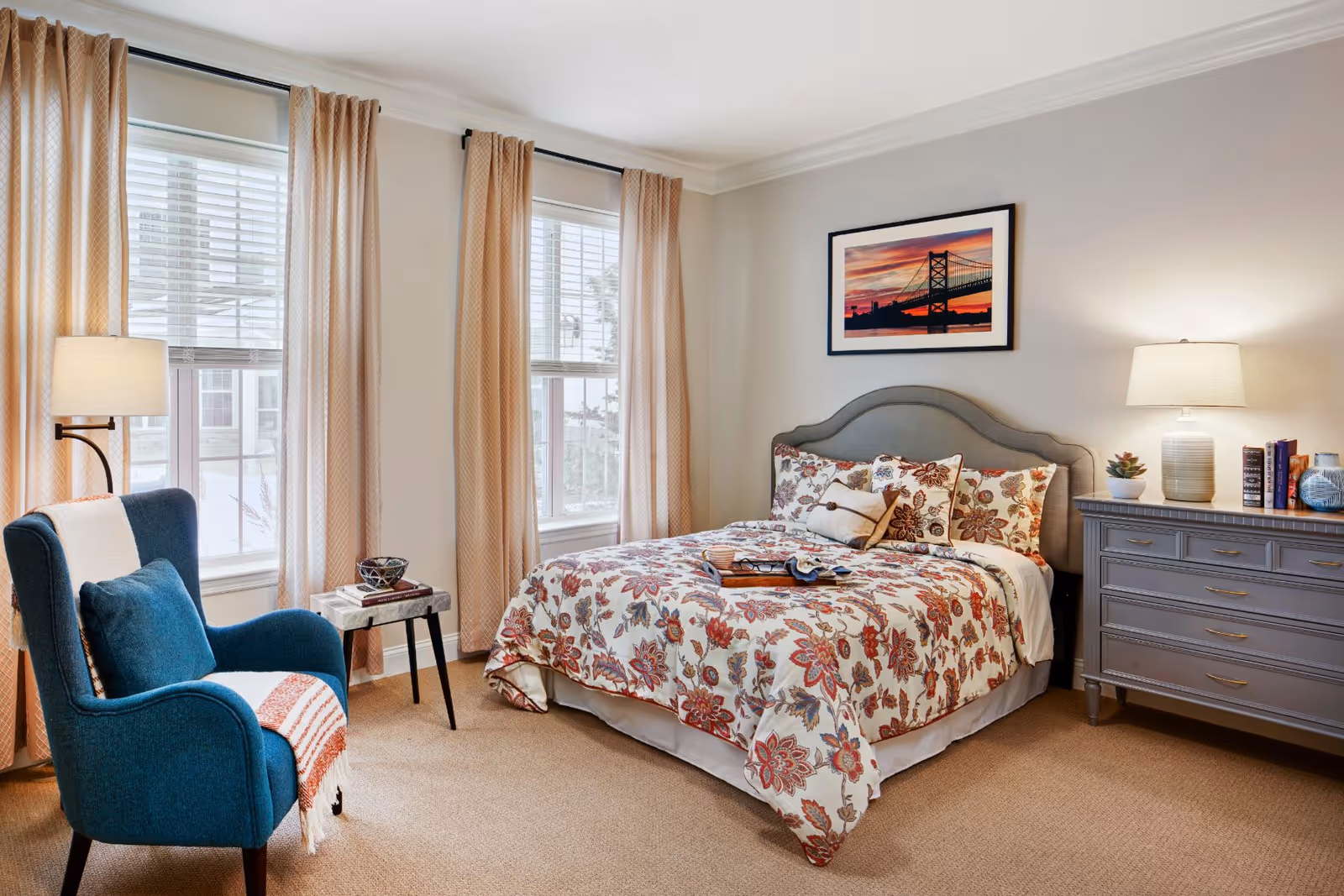A cozy bedroom with a floral patterned bedspread on a bed with a gray upholstered headboard. There are two windows with beige curtains and white blinds, a blue armchair with a white and orange throw blanket, a small side table with a decorative bowl and books, and a gray dresser with a lamp, books, and decorative items. A framed picture of a bridge at sunset hangs on the wall above the bed.