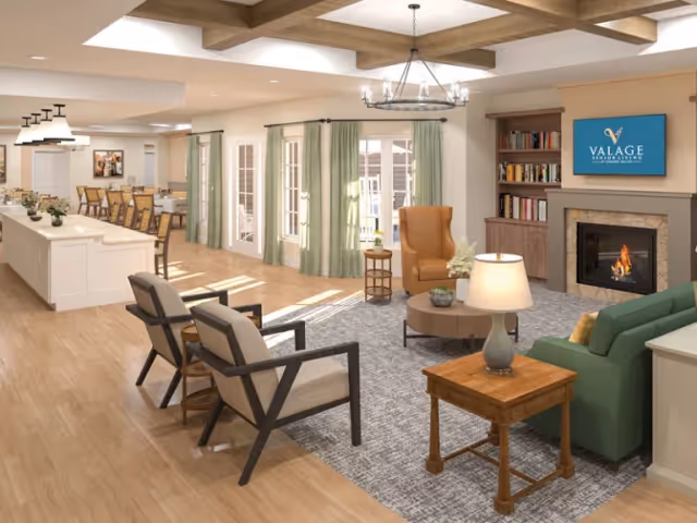 Sunlit open-plan senior living lounge with seating around a fireplace and a TV, and a dining area visible in the background.