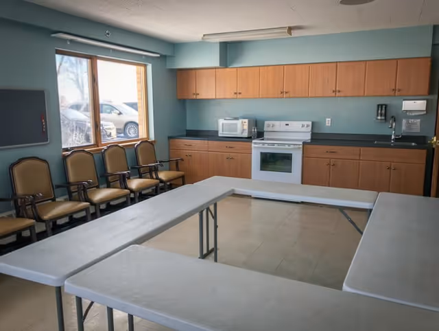 A room with a U-shaped arrangement of white folding tables and a row of chairs along one wall. The room has light blue walls, a window showing parked cars outside, and a kitchenette area with wooden cabinets, a white stove, microwave, and sink.