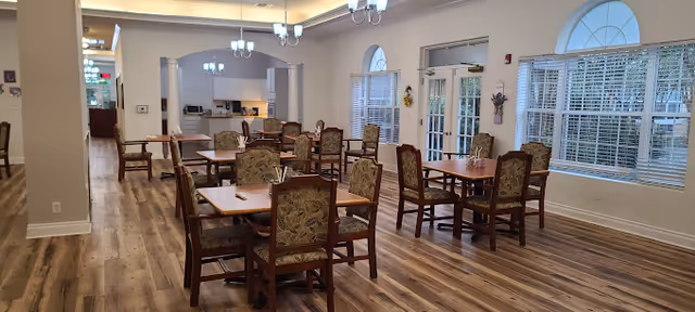 Communal dining room with several wooden tables and upholstered chairs, wood-look flooring, chandeliers, large windows, and a serving area in the back.