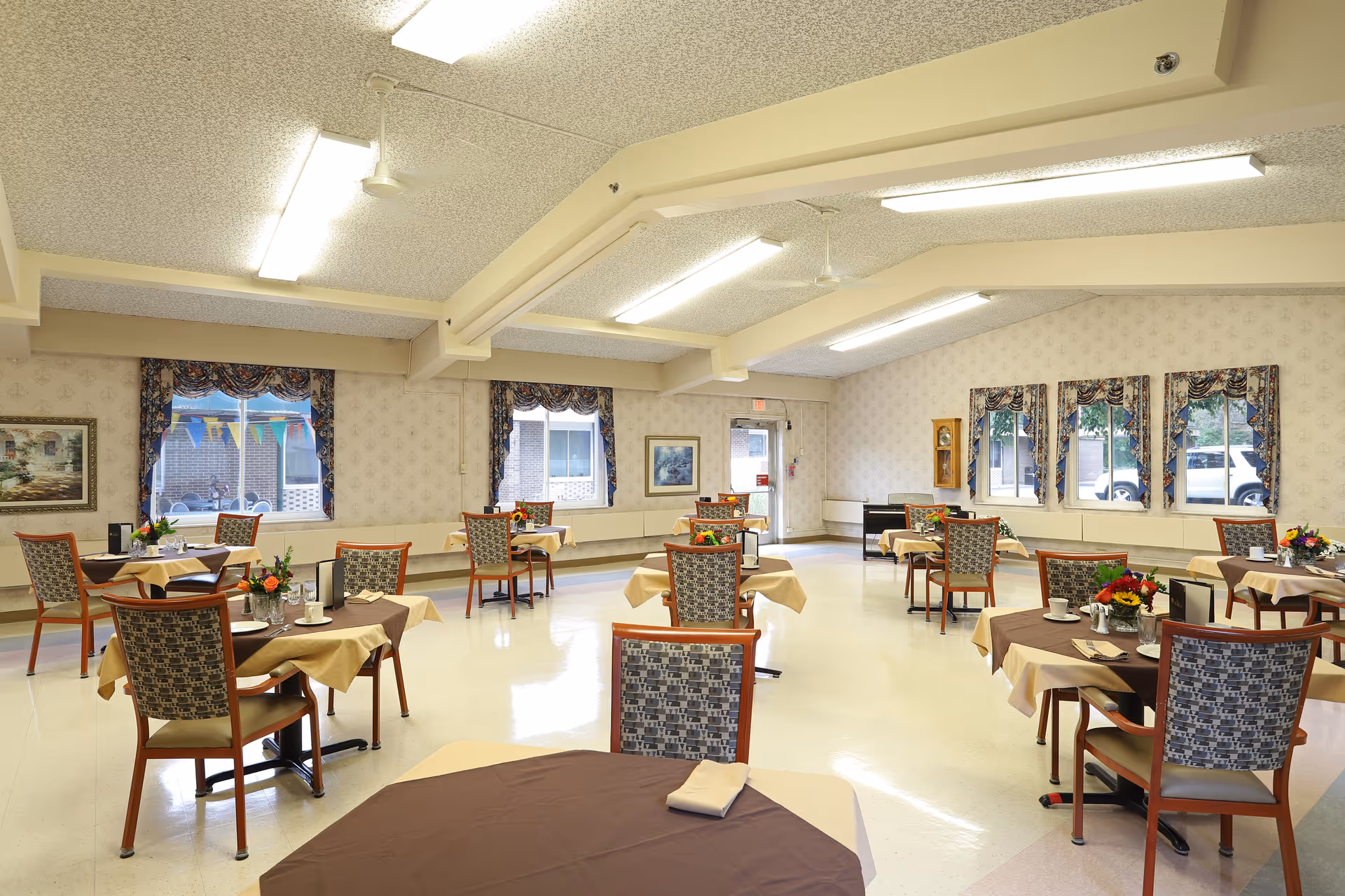 A bright dining room with multiple tables covered with beige and brown tablecloths, each set with cups, napkins, and small flower arrangements. The room has patterned wallpaper, several windows with floral curtains, and fluorescent ceiling lights. Chairs with patterned upholstery are arranged around the tables.