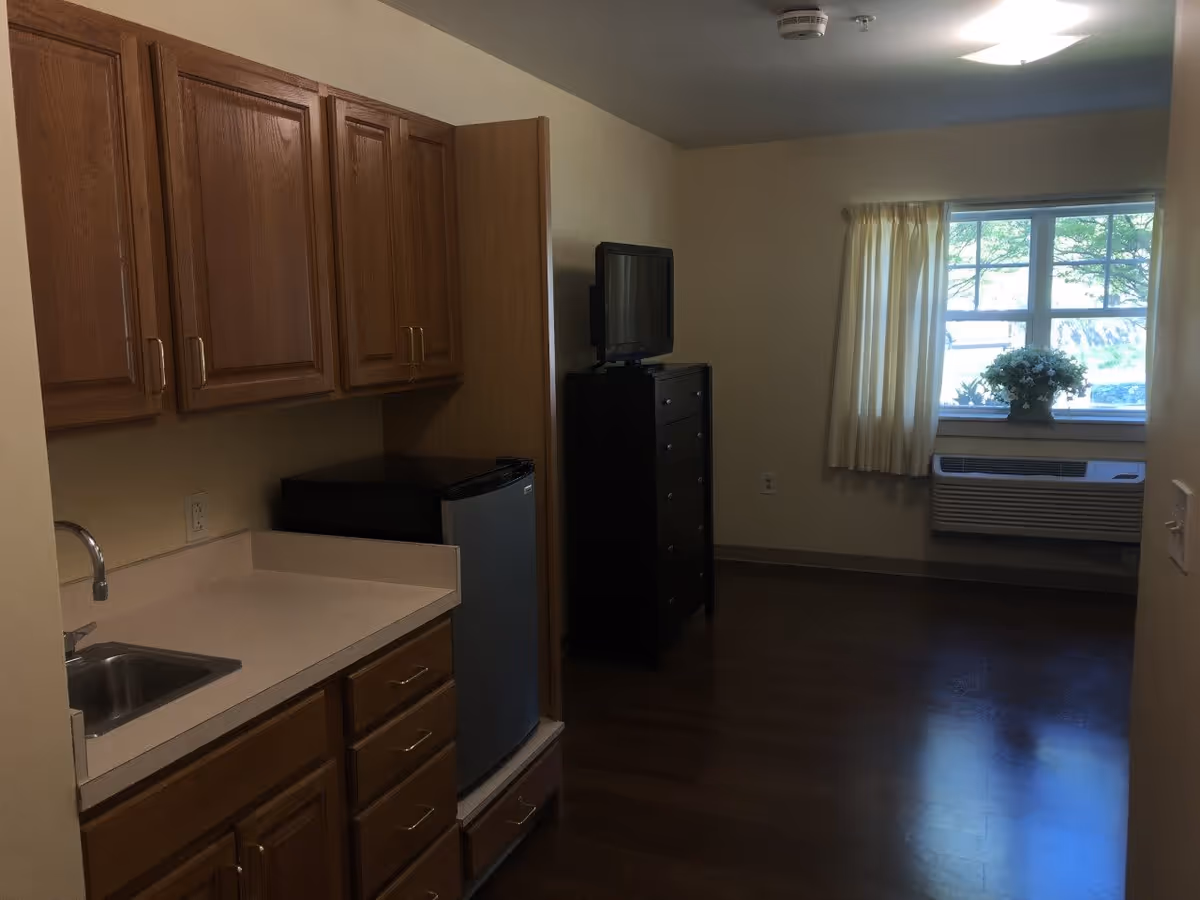 Interior view of a senior living facility room featuring a kitchenette with wooden cabinets, a small sink, and a mini refrigerator. In the background, there is a black dresser with a television on top, a window with cream-colored curtains, and a flower arrangement on the windowsill. The floor is wooden and the walls are painted light beige.
