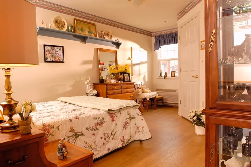 A cozy bedroom with a floral bedspread on the bed, a wooden dresser with a mirror and decorative items, a chair with a teddy bear, a window with blue curtains, and a glass cabinet displaying various items. The room has warm lighting and wooden flooring.